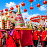 Ilustrasi parade Cap Go Meh dengan peserta berbaju merah mengarak hiasan ritual di bawah deretan lampion merah.