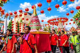 Ilustrasi parade Cap Go Meh dengan peserta berbaju merah mengarak hiasan ritual di bawah deretan lampion merah.