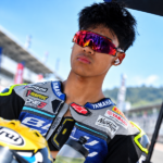Pembalap dengan wearpack Yamaha dan kacamata merah di area sirkuit balap.