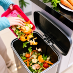 Seseorang membuang sisa makanan berupa kulit buah dan sayuran ke tempat sampah organik di dapur.