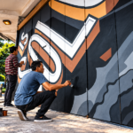 Dua seniman sedang melukis mural grafiti bermotif geometris berwarna hitam, putih, abu-abu, dan cokelat pada dinding bangunan.