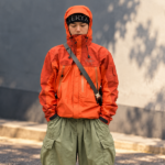 Ilustrasi pria mengenakan jaket outdoor oranye dan celana kargo hijau dengan gaya gorpcore yang menjadi tren fashion 2026.