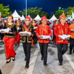 Ilustrasi parade budaya dengan peserta mengenakan busana adat merah, hitam, dan emas sambil membawa wadah tradisional.