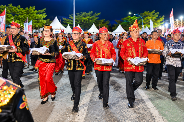 Ilustrasi parade budaya dengan peserta mengenakan busana adat merah, hitam, dan emas sambil membawa wadah tradisional.