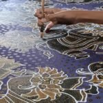 Ilustrasi tangan perajin membuat motif batik menggunakan canting di atas kain bermotif tradisional.