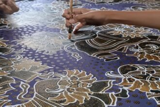 Ilustrasi tangan perajin membuat motif batik menggunakan canting di atas kain bermotif tradisional.