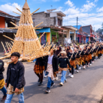 Ilustrasi arak-arakan tradisi masyarakat di jalan desa dengan membawa susunan hasil bumi berbentuk kerucut dalam kegiatan budaya.