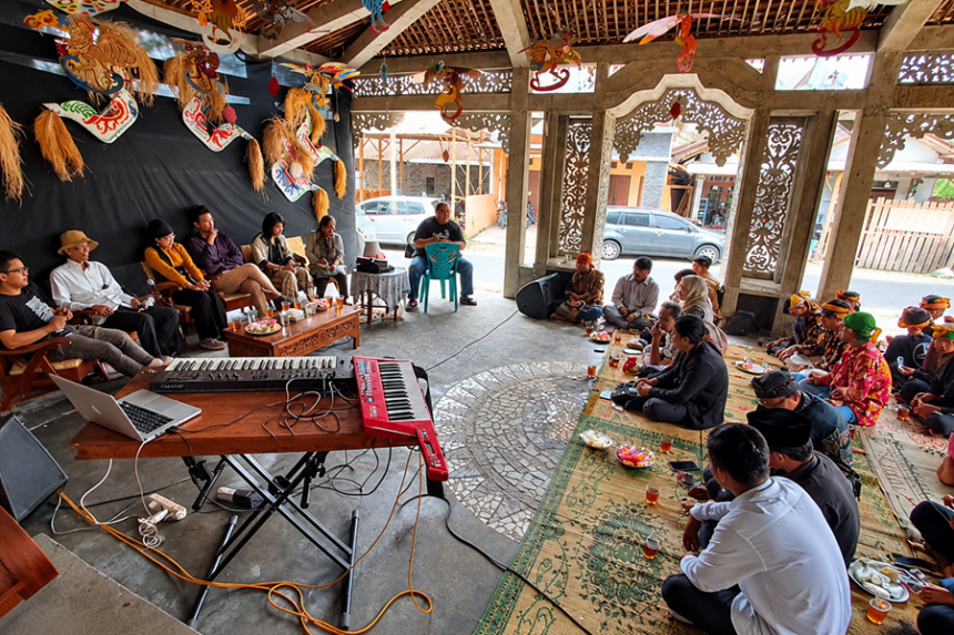 Foto ilustrasi masyarakat berkumpul dan berdiskusi di pendopo tradisional Jawa dengan ornamen budaya dan alat musik.