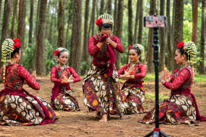 Penari tradisional mengenakan busana batik merah saat menampilkan tarian di tengah hutan pinus.