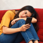 Perempuan duduk di sofa merah sambil menatap ponsel dengan ekspresi sedih