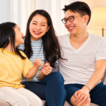 Orang tua dan anak perempuan tertawa bersama di sofa saat menghabiskan waktu bersama keluarga.